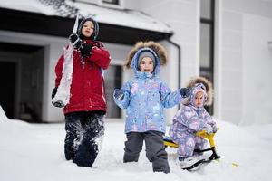 Children play outdoors in snow. Three kids enjoy a sleigh ride. Child sledding in winter against house. photo