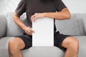 un hombre que sostiene un libro blanco sentado en un sofá gris. foto