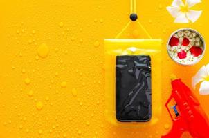 Songkran festival background with waterproof case for smartphone, water gun and flowers in water bowl put on clear mirror that have wet yellow background below. photo
