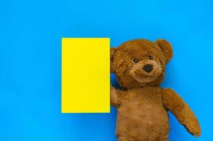 A cute brown teddy bear holding a blank yellow note with blue background for April fools day concept. photo