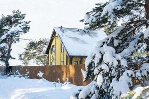 una acogedora casa amarilla en la nieve en invierno en el pueblo está rodeada de pinos. techo cubierto de nieve, tuberías de calefacción y ventilación, ventanas trapezoidales foto