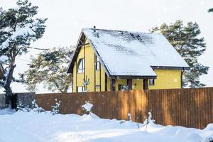 A yellow cozy house in the snow in winter in the village is surrounded by pine trees. Snow-covered roof, heating and ventilation pipes, trapezoid windows photo