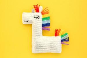 Crochet amigurumi handmade stuffed soft white unicorn toy with rainbow mane on yellow background. Handwork, hobby. Craft diy newborn pregnancy concept. Knitted doll for little baby. Close up flat lay photo