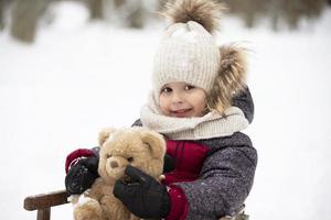 un niño pequeño con ropa abrigada se sienta en un trineo de madera y sostiene un oso de peluche esponjoso. foto