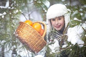 niño feliz con una canasta de naranjas contra el fondo de ramas de abeto cubiertas de nieve. niño con frutas en invierno. foto