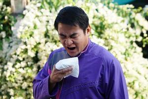 el hombre asiático está estornudando, sosteniendo papel de seda blanco, abre la boca, de pie junto a flores florecientes en el parque. concepto, síntomas de alergia al polen, enfermos, secreción nasal, resfriado. problemas de salud. foto