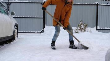 een Mens in winter reinigt sneeuw met een Schep in de werf van een huis in de parkeren kavel. sneeuwval, moeilijk weer conditie, de auto is vertragen, graven omhoog de passage video