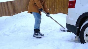 een Mens in winter reinigt sneeuw met een Schep in de werf van een huis in de parkeren kavel. sneeuwval, moeilijk weer conditie, de auto is vertragen, graven omhoog de passage video