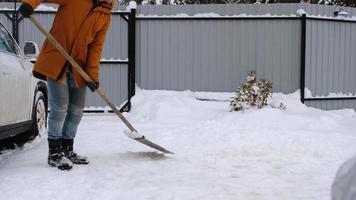 een Mens in winter reinigt sneeuw met een Schep in de werf van een huis in de parkeren kavel. sneeuwval, moeilijk weer conditie, de auto is vertragen, graven omhoog de passage video