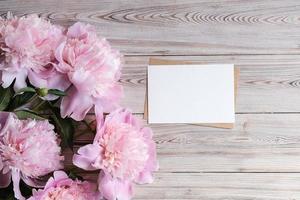 Mock up blank white paper card for text and delicate peonies on a wooden background. Flat lay, top view. Place for text. Greeting card photo