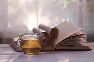 una taza de té verde, un libro abierto sobre una mesa de madera blanca contra el fondo de una ventana a la luz del sol. corazón de las páginas del libro. amor por la lectura. dia de los amantes de los libros foto