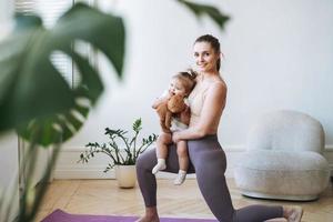 Young woman fit mom with baby girl in her arms doing fitness on mat at home photo