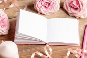 libreta en blanco vacía blanca abierta, rosas suaves, rosadas, velas, pluma, cinta en la mesa de madera vieja. foto