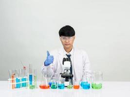 estudiante científico asiático en un laboratorio de mezcla de reactivos en un laboratorio de investigación científica con tubos de ensayo de varios tamaños y microscopios. sobre la mesa en el fondo blanco de laboratorio de química de laboratorio. foto