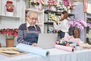 Asian old male florist owner working with a laptop on website for online business, a young beautiful female shopkeeper arranging fresh blossom behind, colorful flower shop store, e-commerce SME. photo