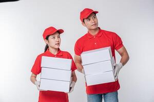 imagen de un joven repartidor feliz con gorra roja en blanco uniforme de camiseta de pie con una caja de cartón blanca vacía aislada en un estudio de fondo gris claro foto