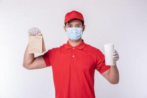 imagen de un joven repartidor feliz con gorra roja en blanco camiseta uniforme mascarilla guantes de pie con un paquete de papel artesanal marrón vacío aislado en un estudio de fondo gris claro foto