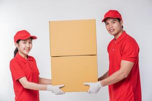 imagen de un joven y feliz repartidor con gorra roja, camiseta en blanco, uniforme de pie con una caja de cartón marrón vacía aislada en un estudio de fondo gris claro foto