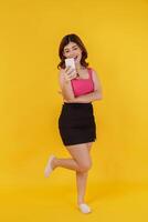 Portrait of Smiling young woman using mobile phone in her hands while standing isolated over yellow background photo