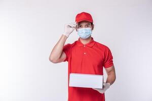 Image of a conscious young delivery man in red cap blank t-shirt uniform face mask gloves standing with empty white cardboard box isolated on light gray background studio photo