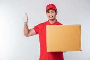 imagen de un joven y feliz repartidor con gorra roja, camiseta en blanco, uniforme de pie con una caja de cartón marrón vacía aislada en un estudio de fondo gris claro foto