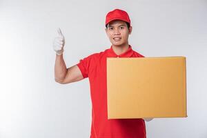 Image of a happy young delivery man in red cap blank t-shirt uniform standing with empty brown cardboard box isolated on light gray background studio photo