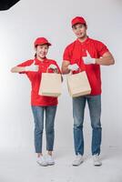 Image of a happy young delivery man in red cap blank t-shirt uniform standing with empty brown craft paper packet isolated on light gray background studio photo