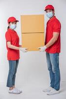 Image of a conscious young delivery man in red cap blank t-shirt uniform face mask gloves standing with empty brown cardboard box isolated on light gray background studio photo