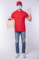 Image of a happy young delivery man in red cap blank t-shirt uniform face mask gloves standing with empty brown craft paper packet isolated on light gray background studio photo