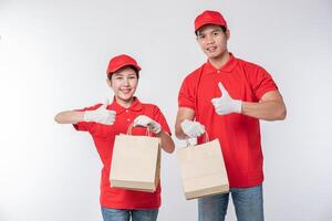 Image of a happy young delivery man in red cap blank t-shirt uniform standing with empty brown craft paper packet isolated on light gray background studio photo