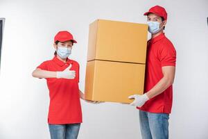 imagen de un joven repartidor consciente con gorra roja en blanco camiseta uniforme guantes de máscara facial de pie con una caja de cartón marrón vacía aislada en un estudio de fondo gris claro foto