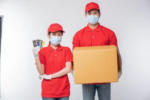 Image of a conscious young delivery man with credit card in red cap blank t-shirt uniform face mask gloves standing with empty brown cardboard box isolated on light gray background studio photo