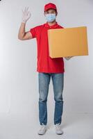 imagen de un joven repartidor consciente con gorra roja en blanco camiseta uniforme guantes de máscara facial de pie con una caja de cartón marrón vacía aislada en un estudio de fondo gris claro foto