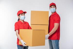 imagen de un joven repartidor consciente con gorra roja en blanco camiseta uniforme guantes de máscara facial de pie con una caja de cartón marrón vacía aislada en un estudio de fondo gris claro foto