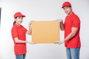 Image of a happy young delivery man in red cap blank t-shirt uniform standing with empty brown cardboard box isolated on light gray background studio photo