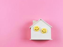 flat lay of wooden model house with two yellow circle smiling faces on it on pink background with copy space. photo