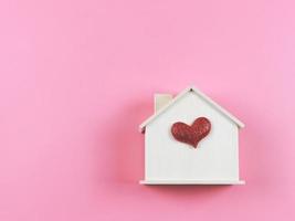 endecha plana de casa modelo de madera con corazón de brillo rojo sobre fondo rosa. casa de ensueño, hogar de amor, relación fuerte, san valentín. foto