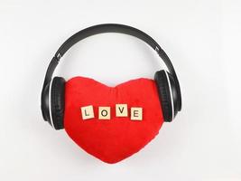 endecha plana de almohada de corazón rojo con letras de madera amor cubierta con auriculares sobre fondo blanco. canciones de amor, podcast o concepto de San Valentín. foto