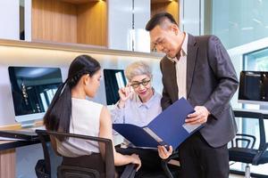 Asian senior business woman giving advice to the new manager and looking at the information regarding sale record while working inside modern style office photo