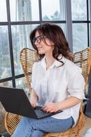 Beautiful businesswoman working on a laptop in a chair near the window. Female freelancer connecting to internet via computer. Blogger or journalist writing new article. photo