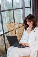 Beautiful businesswoman working on a laptop in a chair near the window. Female freelancer connecting to internet via computer. Blogger or journalist writing new article. photo
