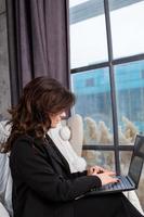 Beautiful businesswoman working on a laptop in a chair near the window. Female freelancer connecting to internet via computer. Blogger or journalist writing new article. photo