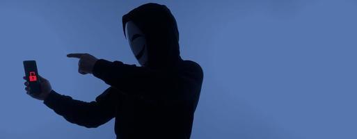 hacker anónimo y mascarilla con smartphone en la mano. hombre con camisa de capucha negra sosteniendo y usando un teléfono móvil con fondo blanco. representan el concepto de piratería informática o robo de datos personales foto