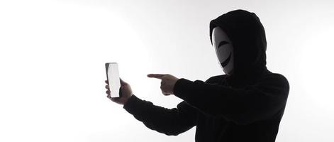 hacker anónimo y mascarilla con smartphone en la mano. hombre con camisa de capucha negra sosteniendo y usando un teléfono móvil con fondo blanco. representan el concepto de piratería informática o robo de datos personales foto