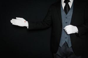 retrato de mayordomo con traje oscuro y guantes blancos de pie con gesto de bienvenida sobre fondo negro. copie el espacio para la industria de servicios y la cortesía profesional. foto