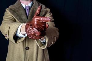 retrato de hombre con abrigo y traje señalando con el dedo el gesto de la pistola sobre fondo negro. espía agente secreto de cine negro. jugando al héroe de acción de detectives. estilo retro y moda vintage foto