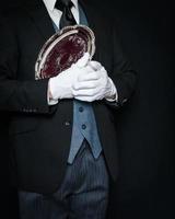 Portrait of Butler in Dark Suit and White Gloves Cradling Silver Serving Tray to His Bosom on Black Background. Professional Service and Hospitality. photo