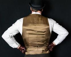 Portrait of Man in Bowler Hat and Vest Standing With Hands on Hips on Black Background. Vintage Fashion and Retro Style. photo