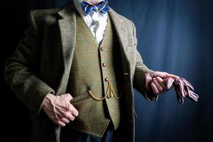 retrato de hombre con traje de tweed sosteniendo un par de guantes de cuero marrón con actitud despreocupada sobre fondo negro. caballero inglés clásico y excéntrico. foto