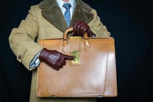 Portrait of Man in Brown Fur Overcoat and Leather Gloves Pointing to Combination on Attache Case on Black Background. Concept of Secret Agent Spy Hero Film Noir. Identity Theft. photo